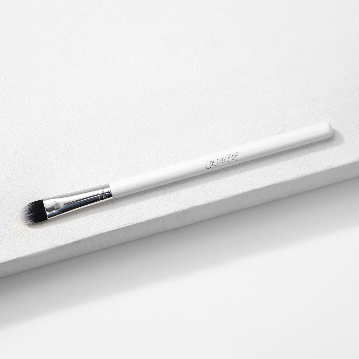 ColourPop Medium Shader Brush with flat oval, dual-toned black-to-white synthetic bristles and a silver ferrule on a white handle, resting on a white textured platform in studio lighting; makeup brush for precise eyeshadow packing and blending.