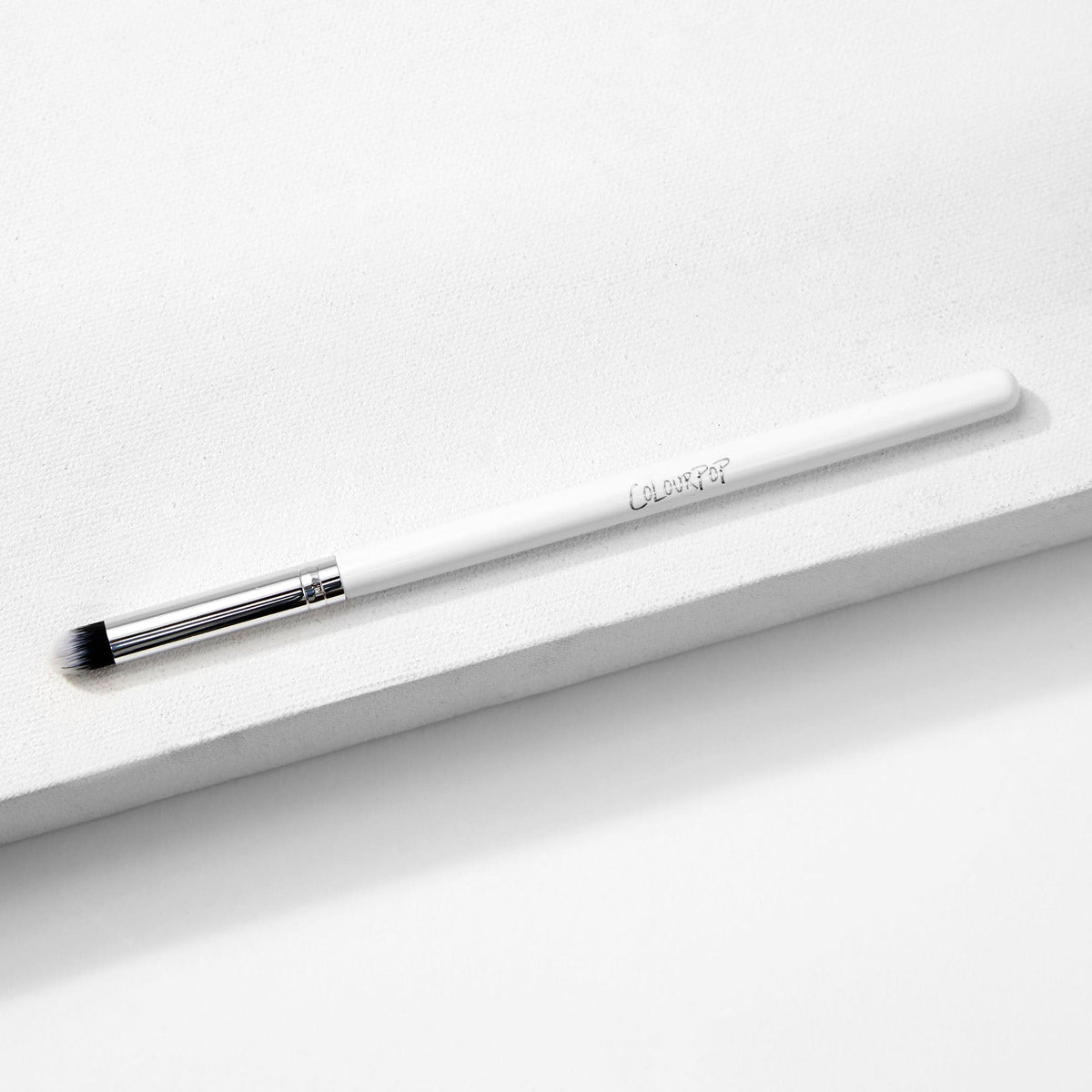 ColourPop Pencil Brush resting diagonally on a white textured block, a makeup brush with a slim white handle and logo, silver ferrule, and small tapered black-and-white bristles for precise eyeshadow and eyeliner smudging.