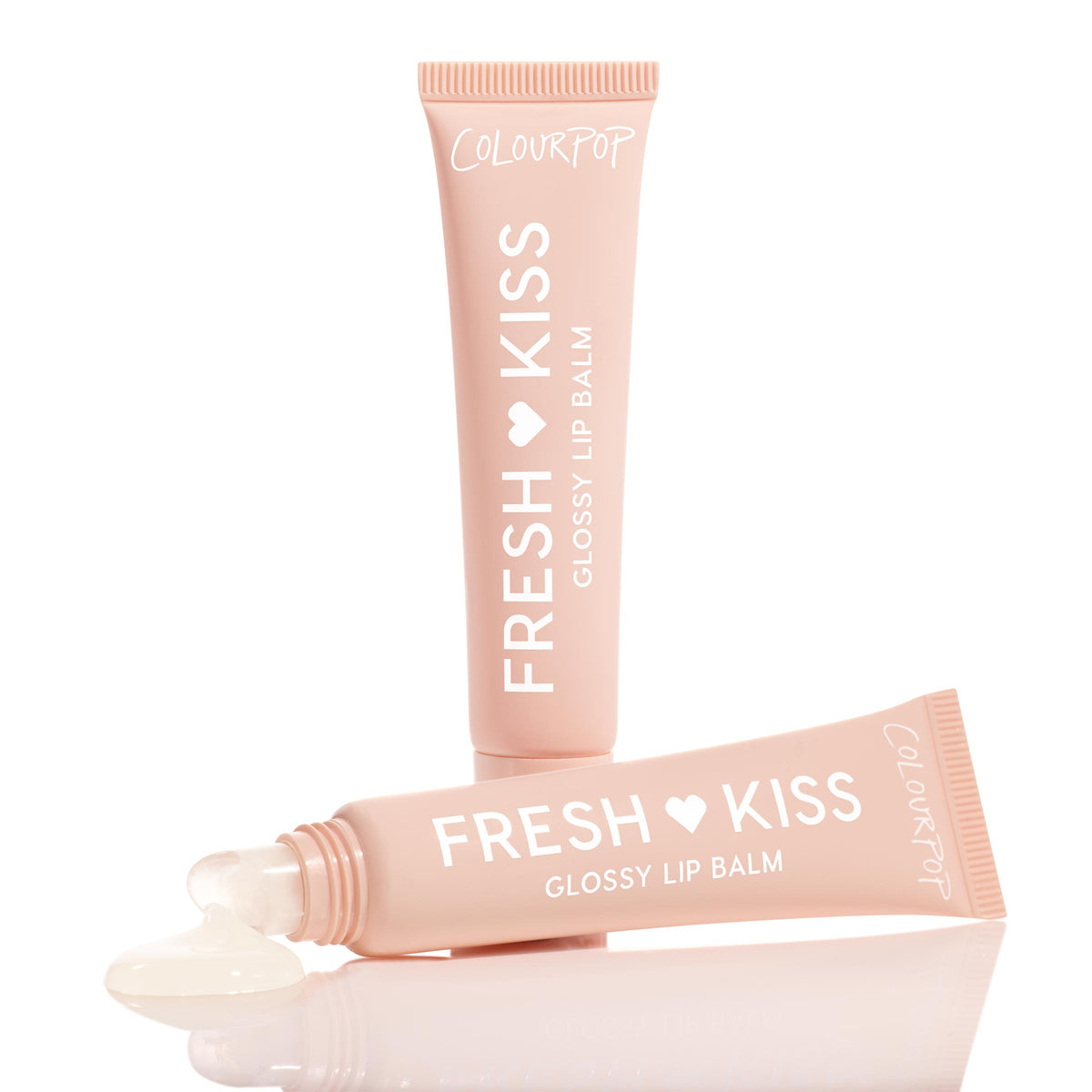 Two soft-pink tubes of ColourPop Fresh Kiss Glossy Balm in Vanilla Cake are arranged on a reflective white surface, one showing a bead of clear, glossy balm pooling from its slanted applicator tip.