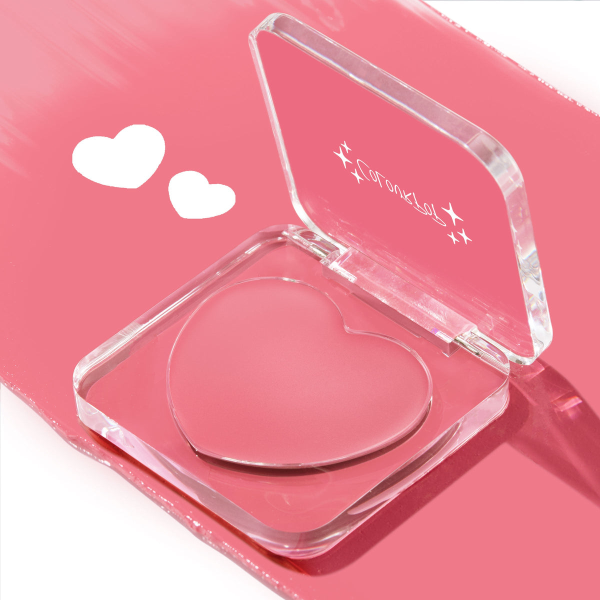 Heart-shaped Lucky U ColourPop Lip and Cheek Balm in an open clear compact, showcasing rosy pink cream; case tilted on a glossy pink background with two white heart doodles, a multitasking lip tint and blush.