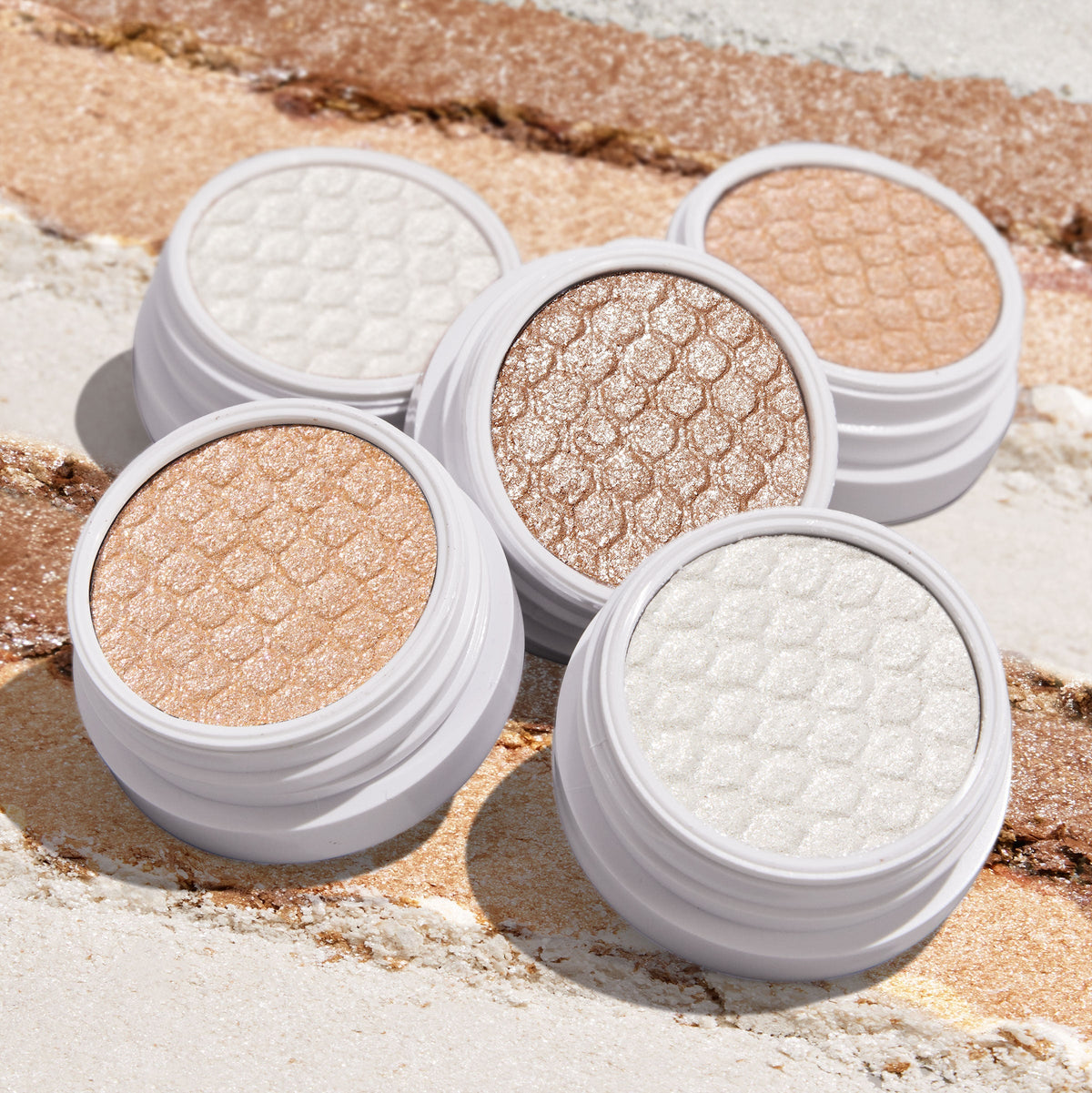 Mighty Morphin' ColourPop Super Shock Shadow opens to show its sparkly metallic, scale-embossed pan in a white twist-top pot, displayed with other neutral shades and crushed swatches across a sandy backdrop.