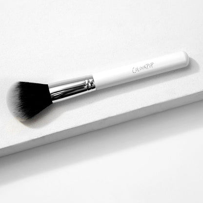 ColourPop Large Powder Brush with fluffy black-to-white bristles rests diagonally on a white textured block, showcasing a chrome ferrule and white handle logo, a soft full-size makeup brush for sweeping on setting or finishing powder.
