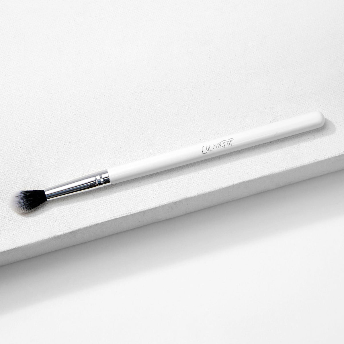 ColourPop Tapered Blending Brush resting diagonally on a white textured surface, featuring a white handle, silver ferrule, and soft tapered black-and-white bristles for seamless eyeshadow blending and crease definition.