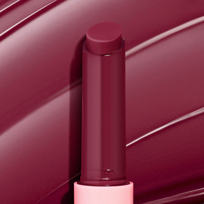 Berry Spritz shade of the ColourPop So Juicy Plumping Gloss Balm with Peptides rising from a light pink tube, the deep berry stick gleaming against a matching glossy swatched backdrop to showcase its smooth, plumping finish.