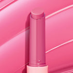 Dolled Up pink twist-up lip balm stick showcasing the ColourPop So Juicy Plumping Gloss Balm with Peptides, standing uncapped to reveal a creamy rosy tint against glossy pink swatch strokes that emphasize shine and a plumping finish.