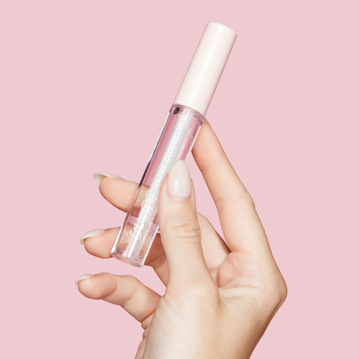 ColourPop Ultra Glossy Lip 20 Carats tube held between fingers, showing clear gloss in a transparent bottle with a pale pink cap and white logo, against a pastel pink background.