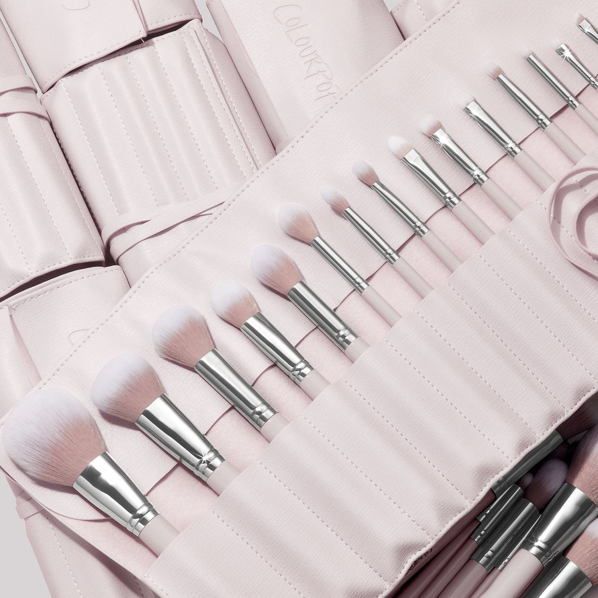 Stone Cold, a ColourPop Makeup Brush Kit, fanned in a blush-pink roll as face and eye brushes slot into stitched pockets, soft white-to-pink bristles and silver ferrules gleaming, with additional matching brush rolls layered behind.