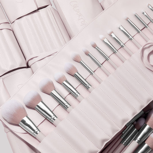 Stone Cold, a ColourPop Makeup Brush Kit, fanned in a blush-pink roll as face and eye brushes slot into stitched pockets, soft white-to-pink bristles and silver ferrules gleaming, with additional matching brush rolls layered behind.