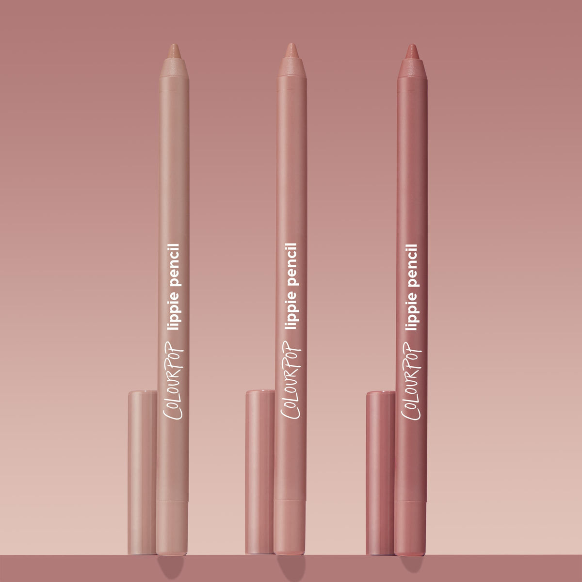 Trio of ColourPop Lippie Pencil Cool Besties standing upright, tips exposed, in light nude, pink nude, and rosy mauve tones with matching caps, arranged on a blush gradient background for a clean product shot.
