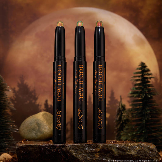 Three Love Triangle ColourPop Shadow Stix stand upright, their shimmery duochrome cream tips glowing green-gold and pink, displayed on a rocky surface with pine trees and a giant full moon backdrop for a moody, twilight woodland vibe.