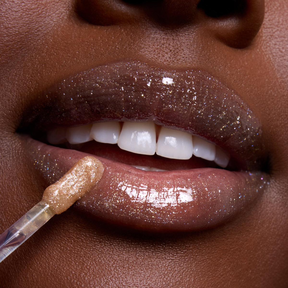 Close-up of glossy, glittering brown lips as a wand applies Ambitious Tiana Ultra Glossy Lip, a shimmery nude Disney Princess lip gloss, highlighting high-shine sparkle and smooth finish on deep skin tone.
