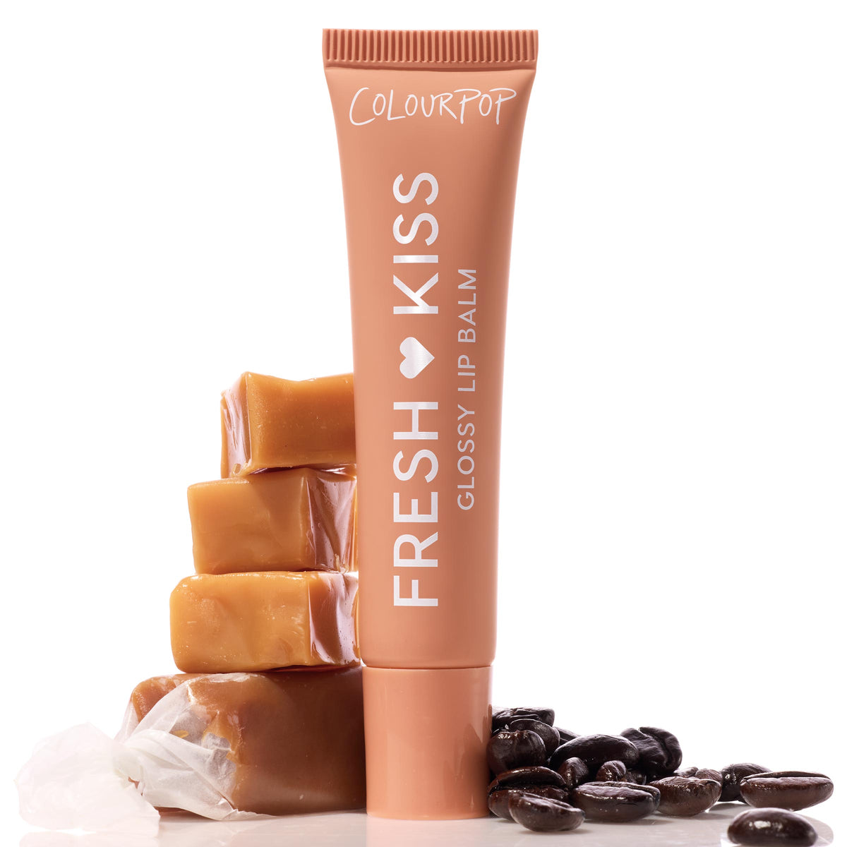 ColourPop Fresh Kiss glossy lip balm in Caramel Latte stands upright in a nude caramel tube beside stacked caramels and coffee beans on a white backdrop, representing the Sweet Treats ColourPop Glossy Lip Balm Trio product packaging.