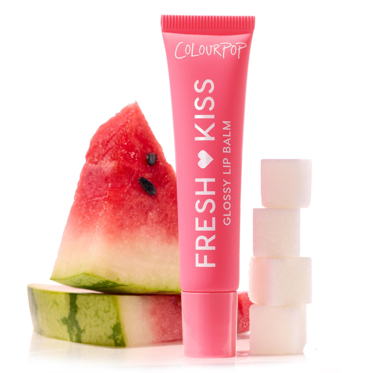 ColourPop Fresh Kiss Glossy Balm in Watermelon Sugar stands upright in a pink squeeze tube, flanked by a seeded watermelon wedge and stacked sugar cubes on a white studio surface, highlighting the fruity, glossy lip balm.