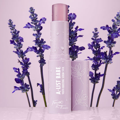 A-List Babe Serum Stick standing uncapped with dewy droplets on a pastel lavender tube, framed by purple lavender sprigs on a glossy pink surface; ColourPop Serum Stick by Fourth Ray Beauty for easy swipe-on face serum.