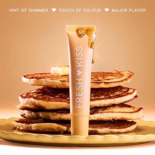 Colourpop Fresh Kiss Glossy Balm in Buttery Pancakes flavor standing upright against a stack of syrup-drizzled pancakes on a yellow plate, styled on a rich burgundy background to showcase this Lip Gloss & Lip Oil hybrid.