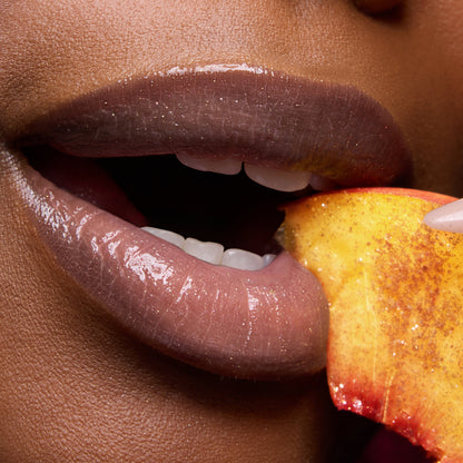 Close-up of shiny, peachy nude lips wearing Colourpop Fresh Kiss Glossy Balm in Peach Mochi, lightly parted as they bite into a juicy peach slice, promoting Lip Gloss & Lip Oil with the tagline “You look good enough to eat.”