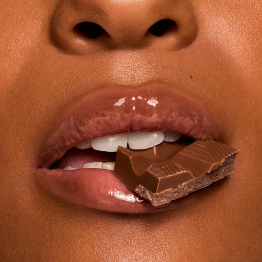 Glossy, cocoa-tinted lips wearing ColourPop Fresh Kiss Glossy Balm in Chocolate Milkshake hold a square of milk chocolate between teeth in a macro close-up, showcasing high-shine, cushy hydration and a creamy nude tint.