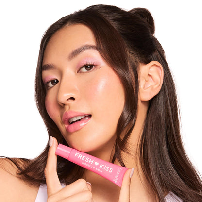Model holds a pink ColourPop Fresh Kiss tube to glossy pink lips, showcasing Fruit Basket, the ColourPop Glossy Lip Balm Trio, in a close-up studio shot on white, emphasizing hydrating shine and a soft pink tint.
