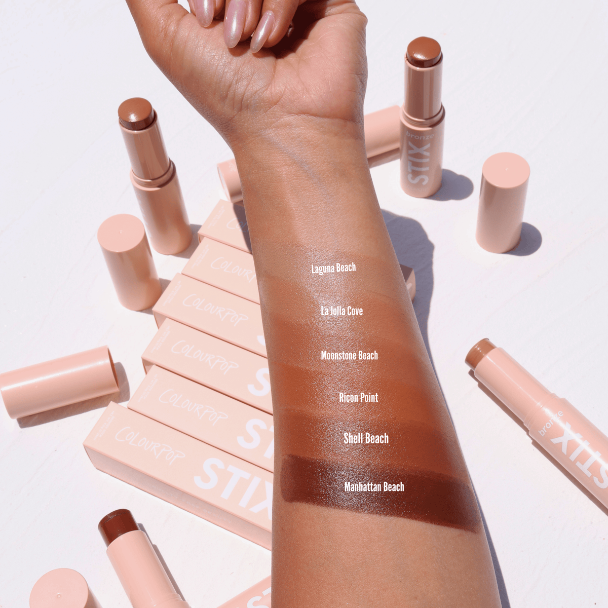 ColourPop Bronzer Stix in Laguna Beach swatched on a forearm - a warm, sun-kissed tan stripe - with additional shade swatches below and nude-pink STIX tubes and boxes scattered on a white surface in bright daylight.