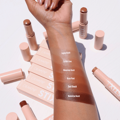 ColourPop Bronzer Stix in Laguna Beach swatched on a forearm - a warm, sun-kissed tan stripe - with additional shade swatches below and nude-pink STIX tubes and boxes scattered on a white surface in bright daylight.