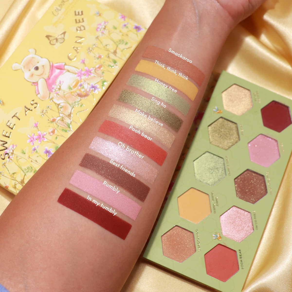 Sweet As Can Bee ColourPop Shadow Palette - open hex-pan eyeshadow palette with warm mattes and shimmering gold, green, pink, and brown shades - swatched on a forearm with names like Smackaroo and Pooh Bear beside Pooh-themed packaging on yellow satin.