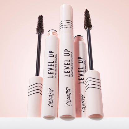 ColourPop Level Up Lengthening Mascara in Brown stands upright, with two open tubes showing slim bristle wands; pale pink packaging with black lines and logos grouped on a white surface against a soft pink backdrop.