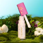 Mother Gothel ColourPop Liquid Highlighter stands open as the doe-foot applicator hovers above the glass tube, revealing a pearly iridescent formula; pink cap with gold flower motif; set on moss with white blooms in a stylized forest under a bright sky.
