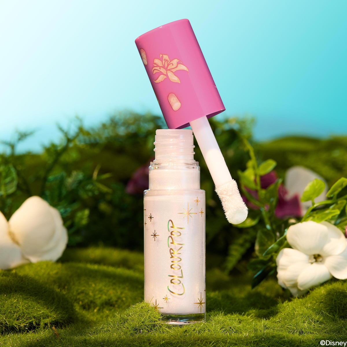 Mother Gothel ColourPop Liquid Highlighter stands open as the doe-foot applicator hovers above the glass tube, revealing a pearly iridescent formula; pink cap with gold flower motif; set on moss with white blooms in a stylized forest under a bright sky.