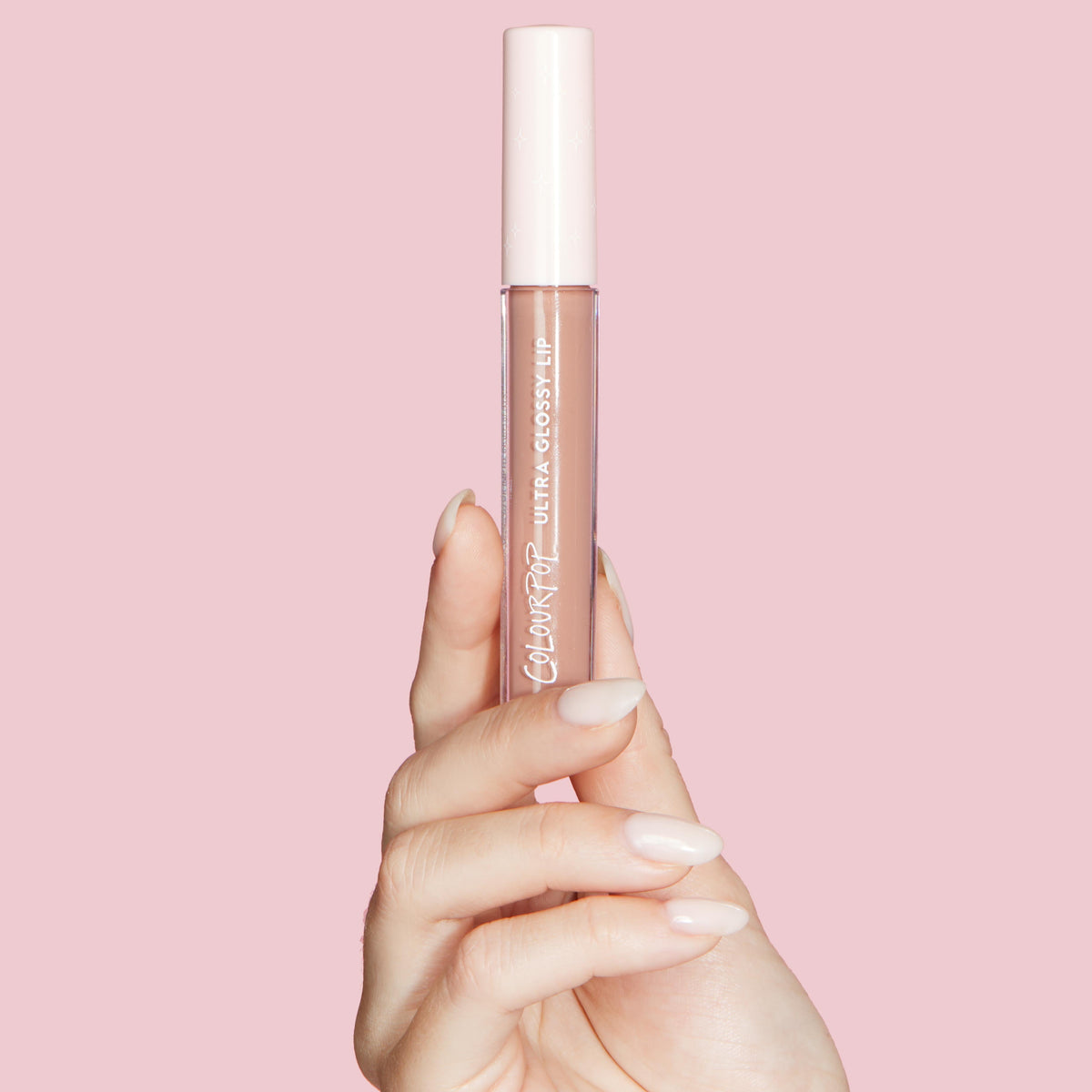 ColourPop Ultra Glossy Lip in Major Throwback held upright by a manicured hand, showcasing a neutral nude-pink gloss in a clear tube with a white cap against a soft pink background.