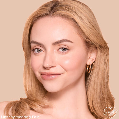 Model wearing ColourPop Pretty Fresh Tinted Foundation Balm in Fair 1, smiling to show a dewy, even complexion; close-up portrait on a beige backdrop with soft nude lips, defined brows, and small gold hoop earrings.