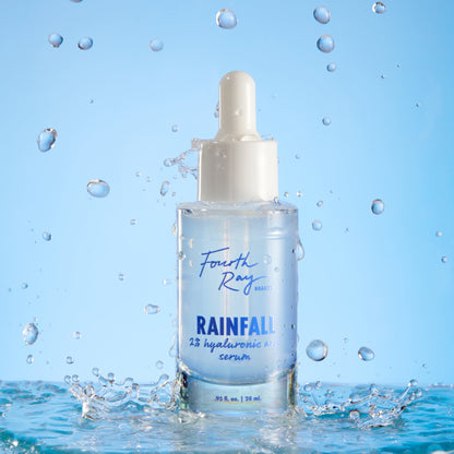 Fourth Ray Beauty Rainfall 2% HA Serum dropper bottle standing upright, catching water splashes against a light-blue backdrop, showcasing its hydrating hyaluronic acid formula; ColourPop Face Serum, 0.95 fl oz/28 mL.