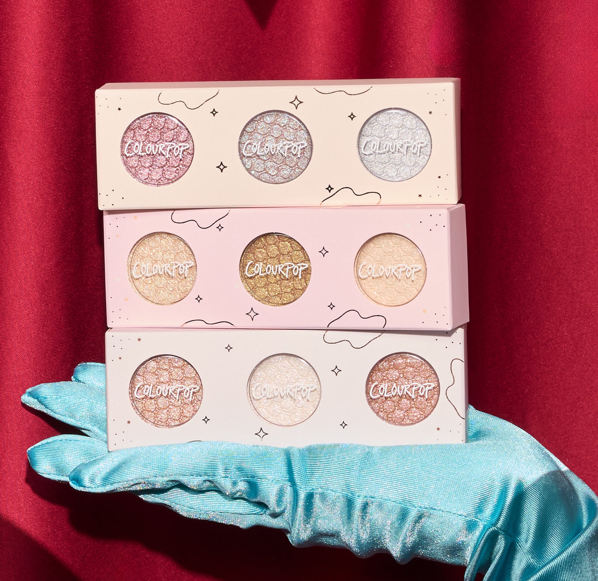 ColourPop Matcha and Mochi Super Shock Shadow trio, three pastel palettes of shimmery eyeshadows stacked on a teal satin glove against a red curtain backdrop, showcasing glittery metallic shades for vibrant eye looks.