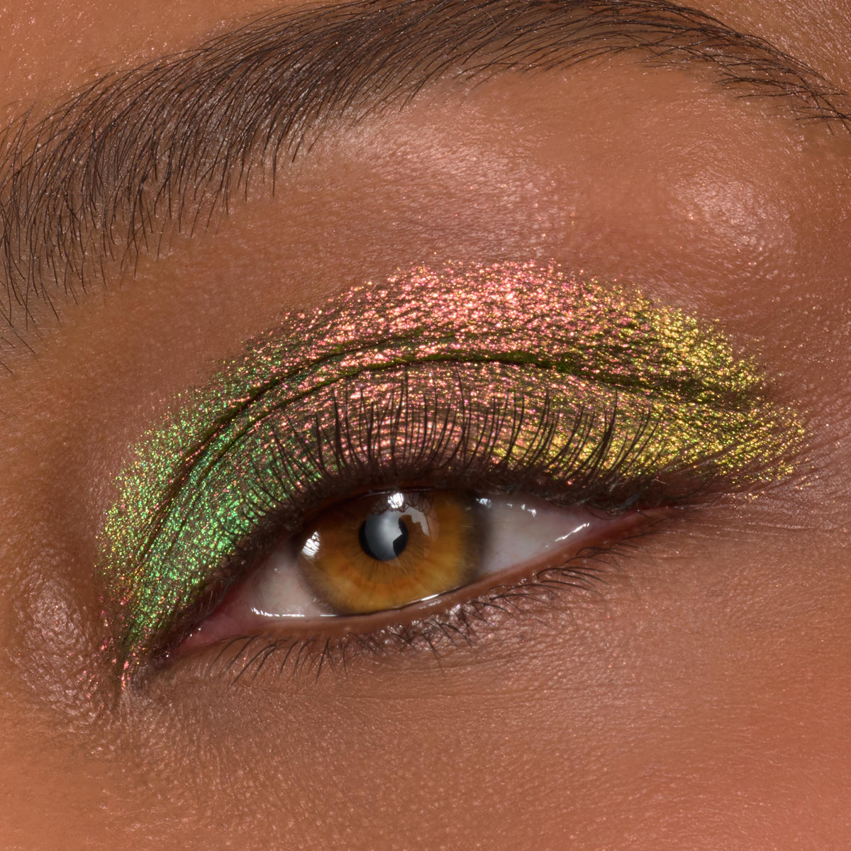Close-up eyelid showcasing It's Always Been Him Shadow Stix, a ColourPop Shadow Stix shade, with multichrome pink, green, and gold metallic shimmer sweeping across the lid and crease on a brown eye, smoothly blended with fine sparkle.
