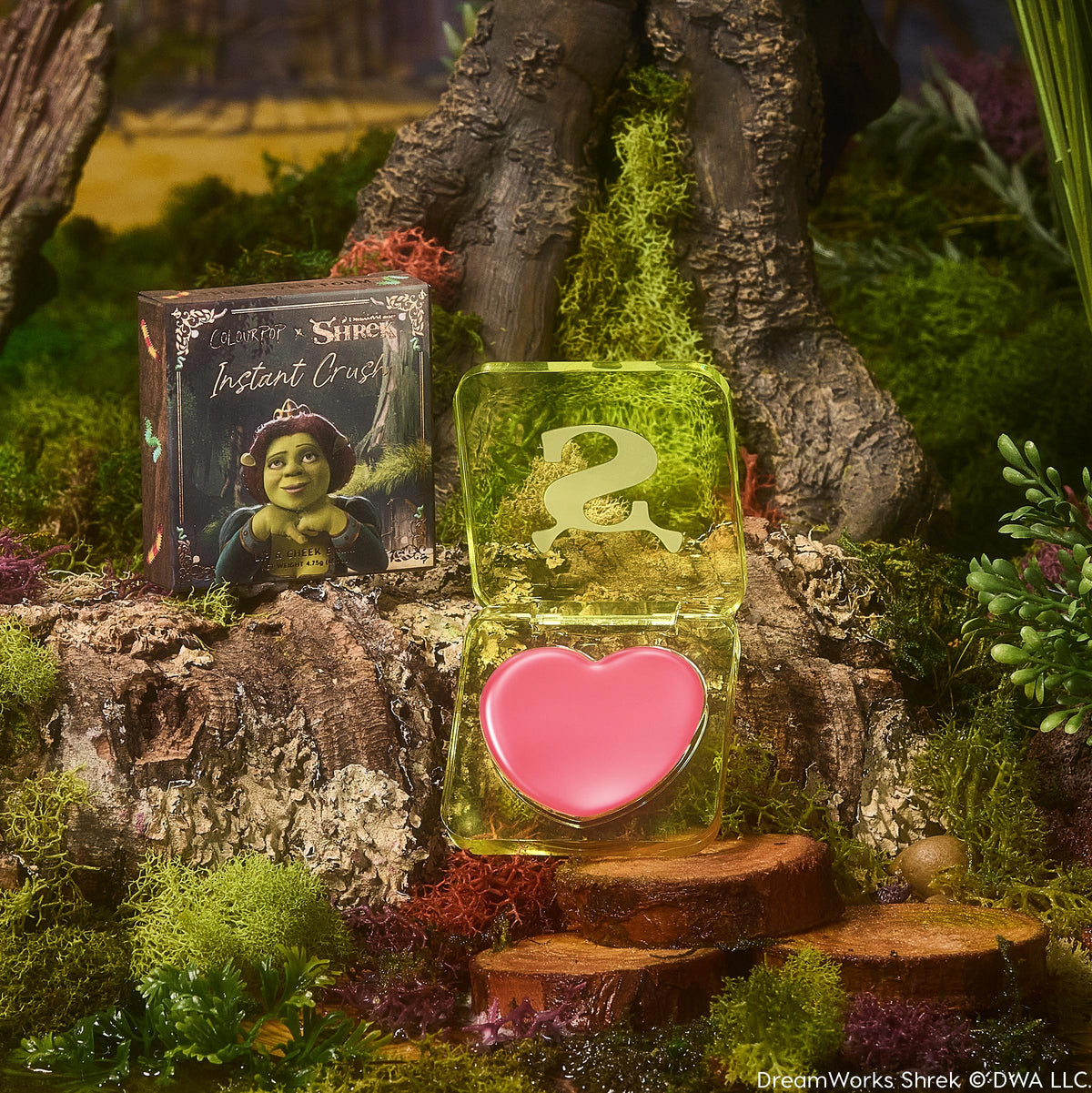 Heart-shaped pink balm sits in an open translucent lime compact with an ogre S logo, beside a box with Princess Fiona artwork, set on a mossy forest log; True Love’s Form ColourPop Lip and Cheek Balm.