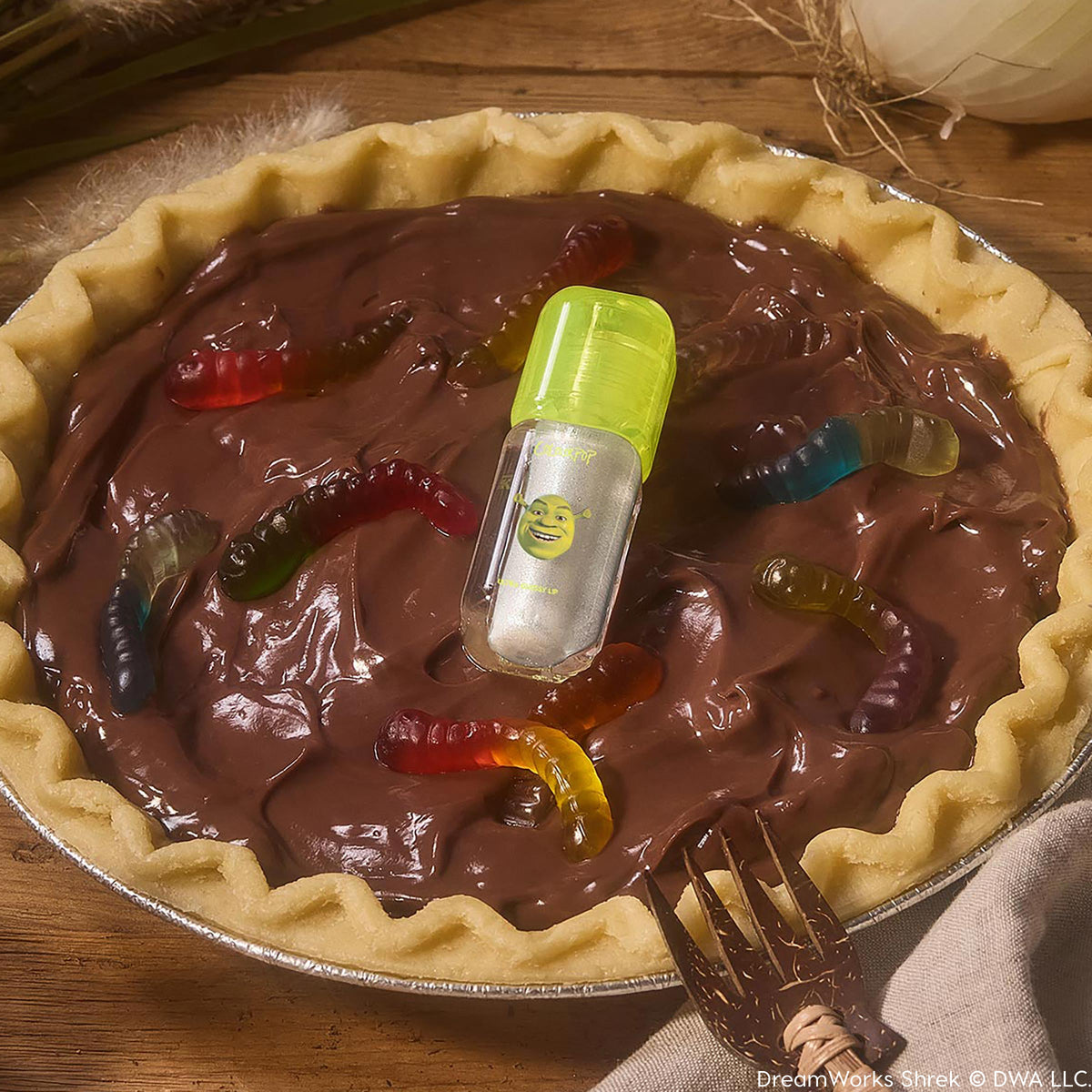 ColourPop Ultra Glossy Lip in Ogre, a shimmering silver gloss in a clear tube with neon green cap and Shrek graphic, rests atop a chocolate mud pie studded with gummy worms on a rustic table, highlighting playful packaging.