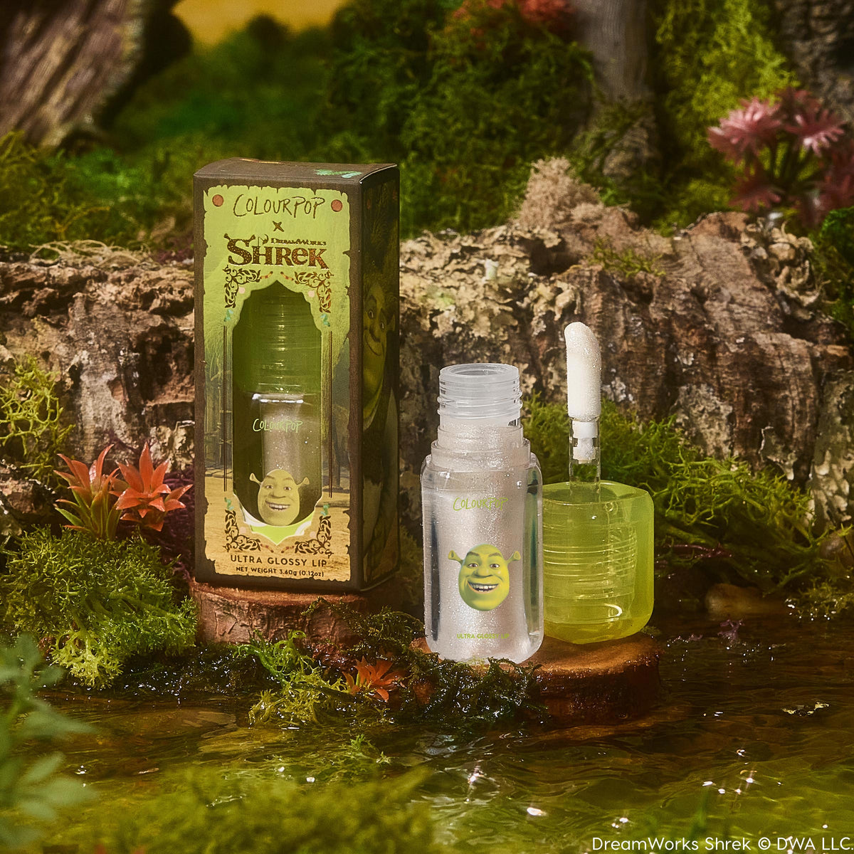 ColourPop Ultra Glossy Lip in Ogre, open with applicator upright beside its box; frosted tube with Shrek face and green cube cap holds a clear, shimmery gloss, staged on a wood slice by water amid moss and flowers in a swamp setting.