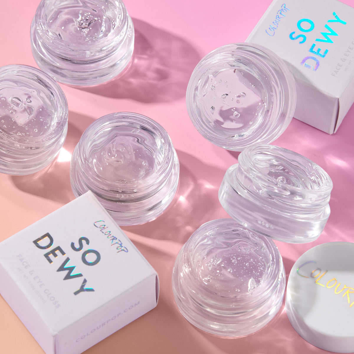 ColourPop Face & Eye Gloss in In The Clear glistening in open glass pots, clear gel catching light, arranged on a pink backdrop beside So Dewy boxes and lids, showcasing the transparent, dewy finish.
