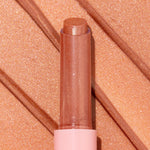 Angel Baby ColourPop So Juicy Plumping Gloss Balm with Peptides extends from a pink tube, the shimmery peach-nude bullet catching light, shown atop matching metallic swatches to highlight its glossy, plumping finish.