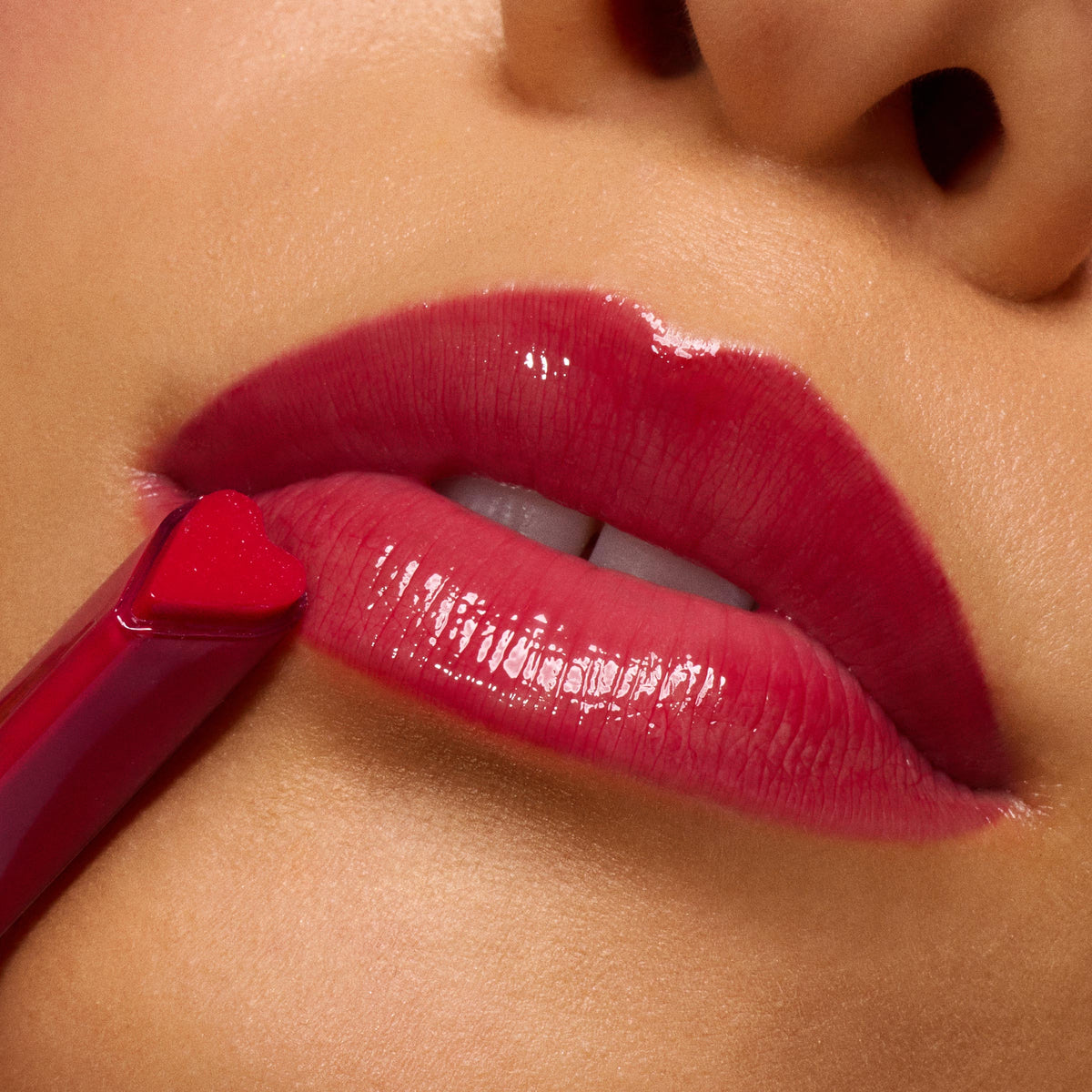 Glossy lips in Spice Level as a heart-shaped applicator applies ColourPop So Juicy Plumping Gloss, delivering a plumped, high-shine cherry-red finish in a tight macro beauty close-up.