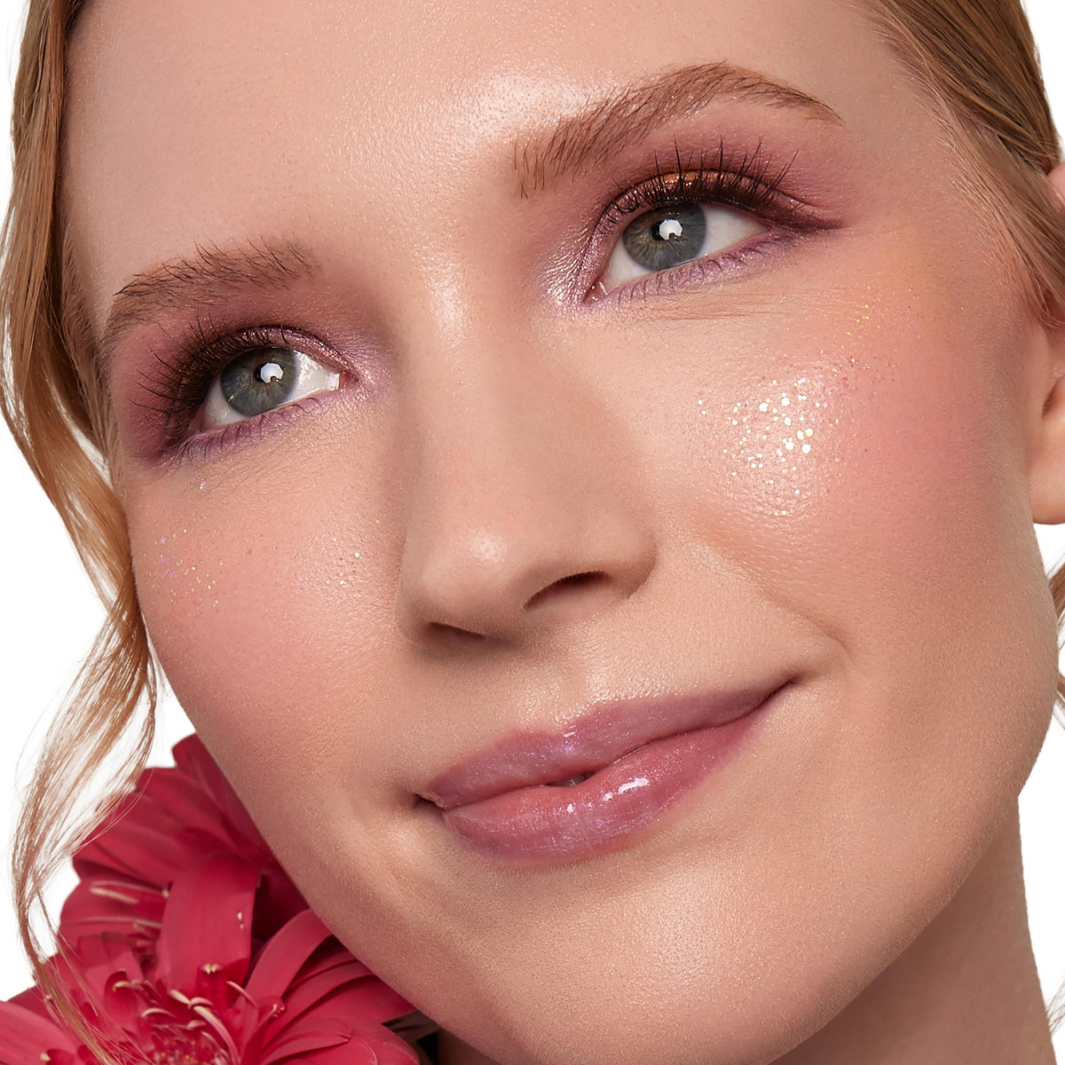 Floating Lights ColourPop Glitter Gel sparkling on a model’s cheeks and under eyes, catching light with iridescent gold flecks; paired with rosy blush, lavender shimmer eyeshadow, glossy lips, and a pink flower accent in a clean studio close-up.
