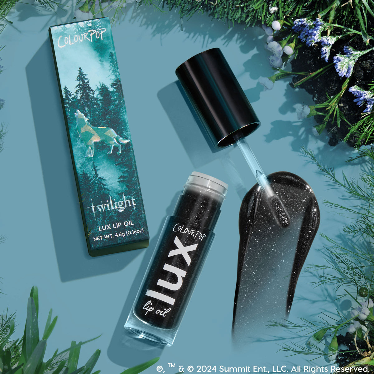 Team Jacob ColourPop Lux Lip Oil shown open with doe-foot applicator and a glossy black, silver-sparkle swatch, beside the Twilight-themed box with wolf and forest art, 4.6g (0.16oz), on a teal flat lay with pine and wildflower accents.