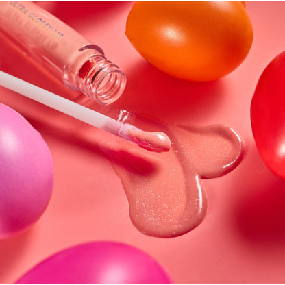 One Kiss Ultra Glossy Lip applicator swipes peachy‑pink shimmer gloss into a heart-shaped puddle beside its open tube on a coral surface with colorful rounded props; ColourPop Ultra Glossy Lip shows a high-shine, cushiony finish.