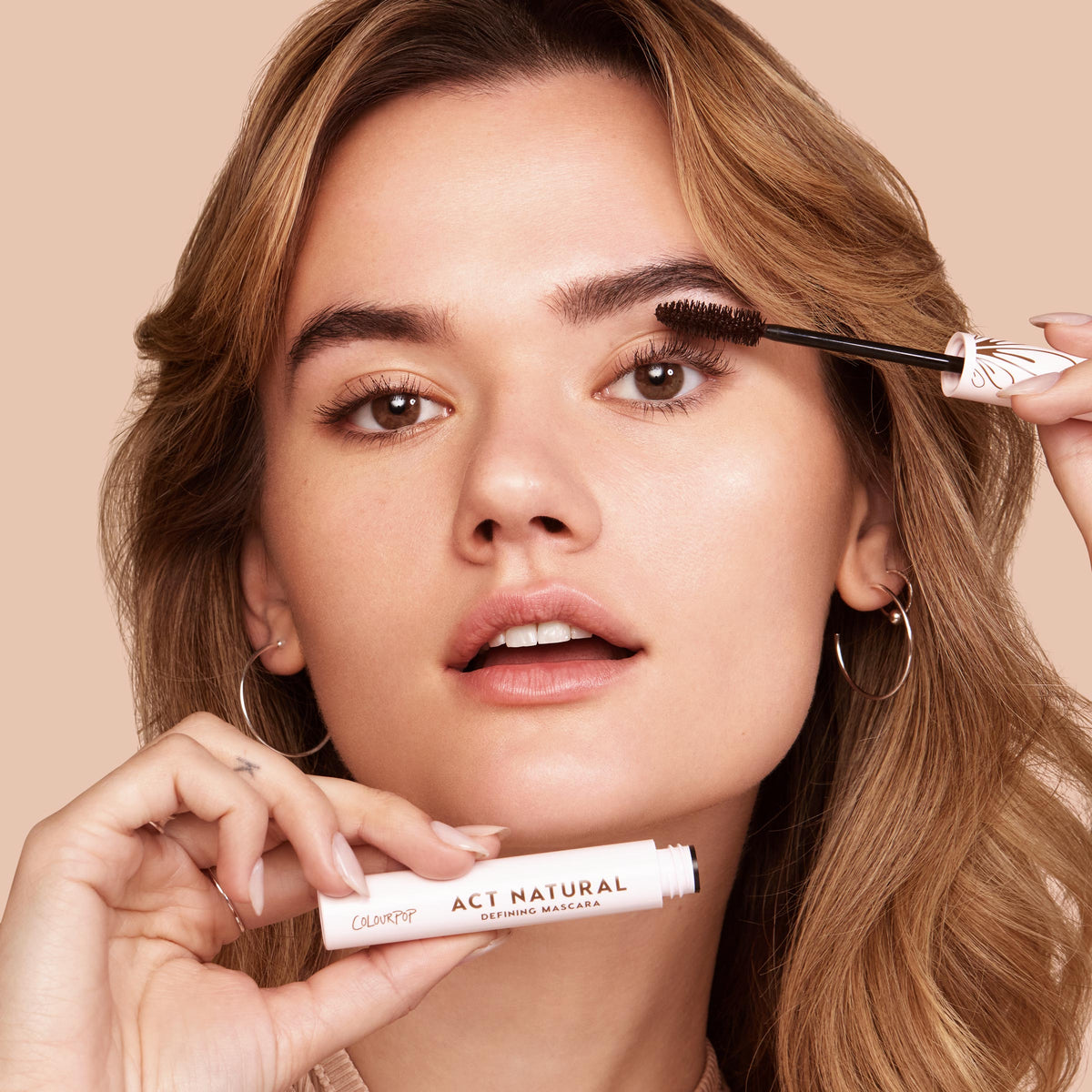 ColourPop Act Natural Mascara in Black defining lashes - model sweeps the black formula with a thick bristle brush while holding the light-pink tube labeled Act Natural, close-up against a neutral beige background.