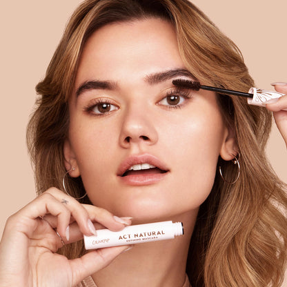 ColourPop Act Natural Mascara in Black defining lashes - model sweeps the black formula with a thick bristle brush while holding the light-pink tube labeled Act Natural, close-up against a neutral beige background.