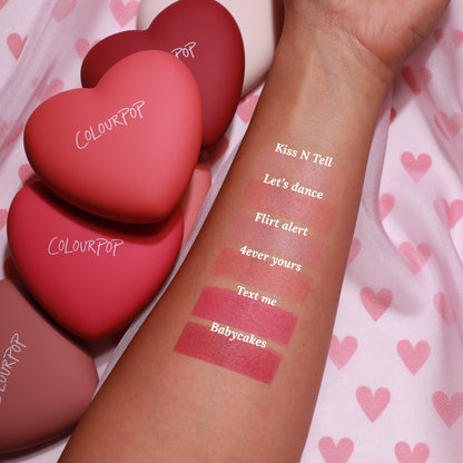 ColourPop Kiss 'N Tell pressed powder blush swatched on a forearm, showing a soft rosy hue beside labeled shade swatches, with matte heart-shaped ColourPop compacts in pink and red resting on a pink heart-print fabric.