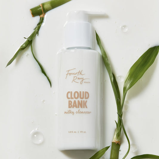 Cloud Bank Milky Cleanser bottle by Fourth Ray Beauty rests on a glossy, milk-like surface with water droplets, framed by green bamboo leaves; white pump-top 5.8 fl oz (171 ml) packaging, presented as a gentle ColourPop Face Cleanser.