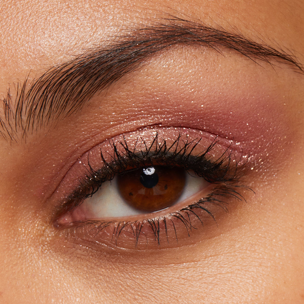 ColourPop Roy G. Biv Super Shock Shadow glitters on the eyelid, delivering a rosy pink base with gold sparkle and a creamy metallic finish, shown in a macro close-up of a brown eye with lashes and brow.